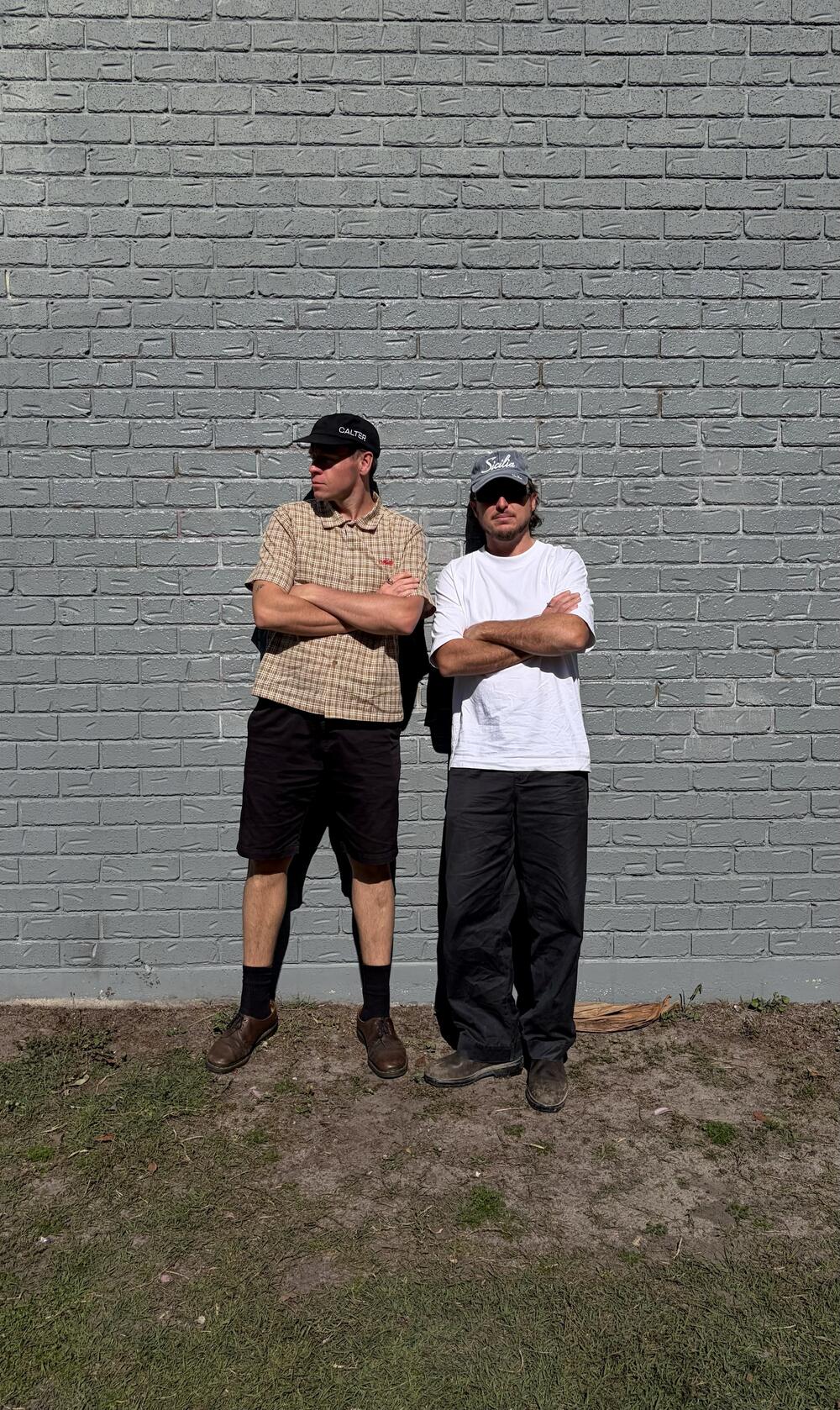 Bali and Benny Benson leaning on a brick wall, arms crossed
